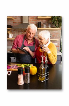 Happy caucasian senior couple preparing vegetables, drinking wine and using tablet in kitchen