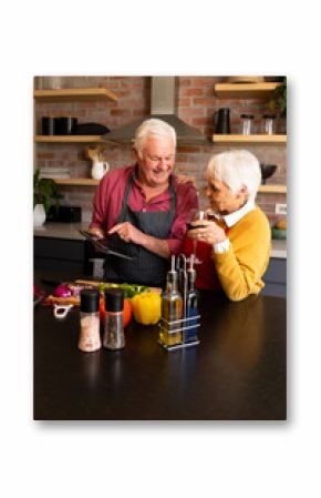 Happy caucasian senior couple preparing vegetables, drinking wine and using tablet, copy space