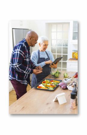 Diverse senior couple preparing meal using tablet in kitchen