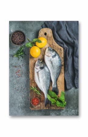 Fresh uncooked sea bream or dorado fish with lemon, herbs and spices in bowls on rustic wooden board over grey concrete background, top view. Healthy, dieting, clean eating concept