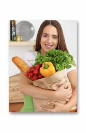 Young caucasian woman in a green apron is holding paper bag full of vegetables and fruits while smiling in kitchen