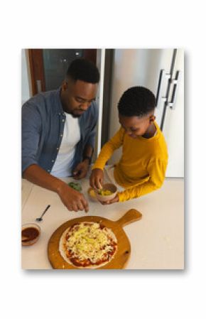 African American father and son making pizza in modern kitchen with pizza board and toppings bowl