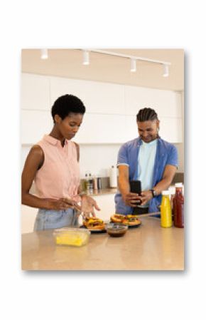 Diverse friends preparing cheeseburgers at island in home kitchen, with smartphone capturing moment