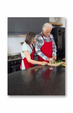 Senior couple in kitchen preparing vegetables together, wearing matching red aprons, at home