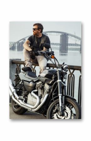 Redhead biker with beard in leather jacket sitting on railings near motorbike on bridge