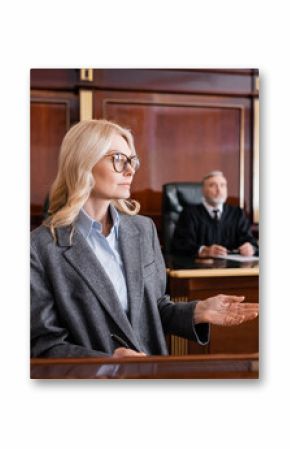 blonde advocate pointing with hand while talking in court near judge on background