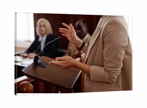 Cropped shot of young female witness in beige blazer standing by tribune with microphone and testifying in front of jury, judge and protection side