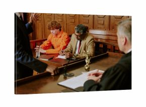Caucasian teenage boy with curly red hair sitting in courtroom wearing orange jumpsuit, accompanied by middle aged biracial man writing notes, judge and attorney present during trial