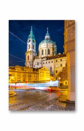 A blurred tram moves through Lesser Town Square in Prague at night, with the stunning architecture of St. Nicholas Church illuminated by streetlights.