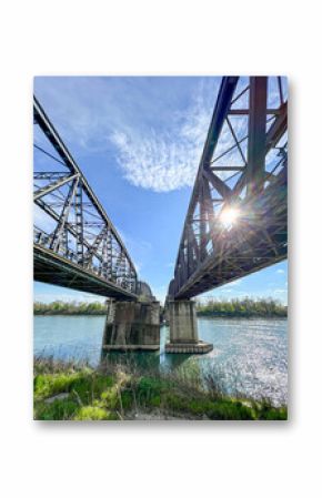 A low-angle view of two robust metal truss bridges spanning the Po River near Cremona, with bright sunlight glinting off the water and a clear blue sky.