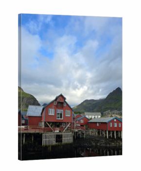 Colorful red cabins perched over the waterfront in A Lofoten, Norway, are framed by dramatic mountains and cloudy skies, creating a picturesque view that captures the essence of Norwegian coastal life