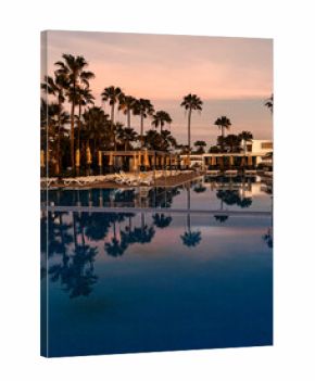 Palm trees and sun loungers reflected in still resort pool at sunset with soft pink sky.