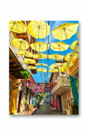 Bright yellow umbrellas and international flags hang above a narrow pedestrian street lined with colorful colonial buildings in Cartagena, Colombia. The lively decorations create a festive atmosphere