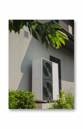 Vertical outdoor heat pump unit installed next to a house wall, surrounded by green shrubs in a modern landscaped garden
