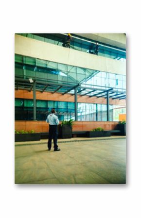Security guard observes window washers working high on a modern Bengaluru building.  Low angle composition emphasizes height.