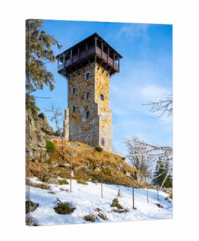 A historic lookout tower stands on Wysoki Kamien in the Jizera Mountains of Poland, surrounded by snow-covered ground, tall trees, and a clear blue sky, inviting visitors year-round.
