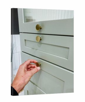 Hand adjusting brass cabinet knob over mint drawer, gentle grip suggesting human interaction, warm bathroom tile backdrop, closeup detail conveying tactile lifestyle moment and modern