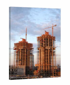 Two tall buildings under construction with cranes in the background. The sky is cloudy and the sun is setting