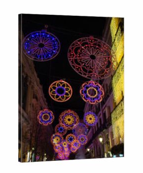 Colorful festive street christmas lights hanging above a city street at night, forming circular patterns and illuminating historic buildings, creating a vibrant holiday atmosphere with urban