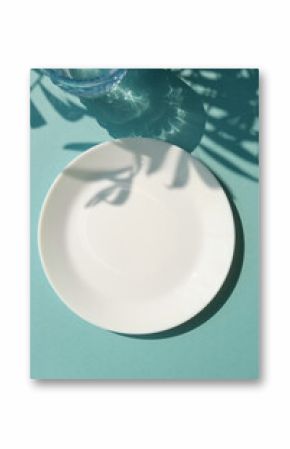 Empty white plate with glass of water on the blue background with palm leaves shadows. Summer table settings. Copy space
