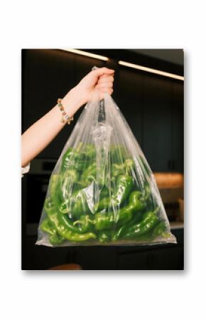Womans Hand Holding a Transparent Bag Filled with Fresh Green Peppers Against a Dark Kitchen Background.