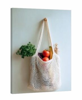 Reusable cotton mesh bag filled with fresh produce including tomatoes, bananas, and parsley hanging on white wall, zero waste lifestyle concept.
