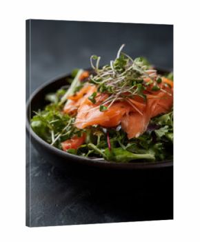 Vibrant salad with fresh green leaves, tomatoes, and smoked salmon on microgreens in black bowl against dark background
