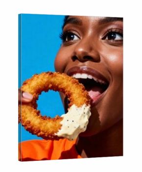 A young woman with dark skin takes a bite of a crispy onion ring dipped in sauce
