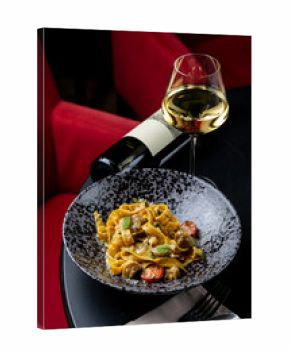 A beautifully plated pasta dish garnished with fresh ingredients sits on a stylish black plate beside a shimmering glass of white wine. The warm ambiance creates a perfect dining experience