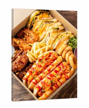 A high-angle close-up shot of a fast-food delivery box filled with various items: hot dogs, french fries, onion rings, fried chicken, and sandwiches on a wooden table.