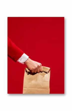 Hand in red suit holding kraft paper bag over red checkered table against vivid red background. Concept of food, emotions, pop art photography, takeaway, delivery services. Vertical layout