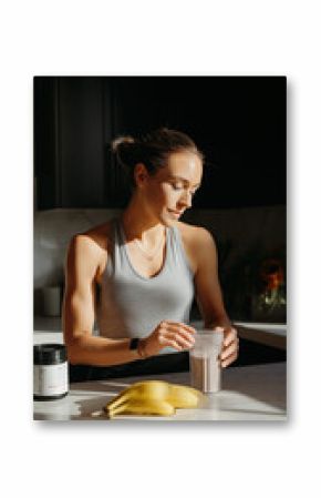 A white woman in her 30s prepares a protein shake in a stylish kitchen, with bananas and a supplement jar on the counter, showcasing a healthy lifestyle and focus on fitness