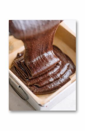 Close-up image of rich, thick chocolate batter being poured into a square baking tin lined with parchment paper. The glossy texture and fudgy consistency suggest preparation for brownies or a moist. 