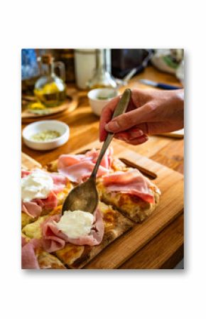 Cooking Pinsa Romana with mozzarella and burrata cheese, boiled ham, tomatoes and olives on wooden kitchen table, close-up