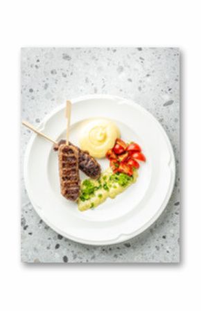 Grilled meat skewers with mashed potatoes, cherry tomatoes, and creamy herb sauce on a white plate over a gray speckled surface