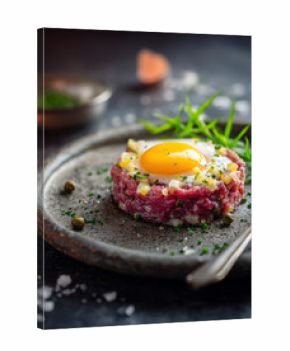 Close-up of steak tartare topped with raw egg yolk, served with diced onion and capers on rustic plate, elegant food styling.