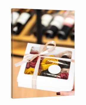 Gift box with colorful pasta nests tied with ribbon, held by hand with red nails. Wine bottles in background. Gourmet food presentation in elegant packaging