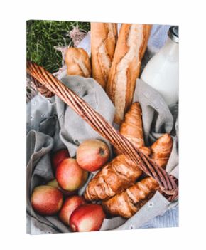 A wicker basket filled with fresh apples, croissants, baguettes, and a bottle of milk.