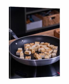 Cubes of golden-brown tofu are sizzling in a non-stick frying pan on a modern stovetop, showcasing the cooking process and vibrant kitchen environment