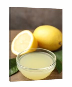 Lemon juice in bowl, leaves and fresh lemons on wooden table, closeup