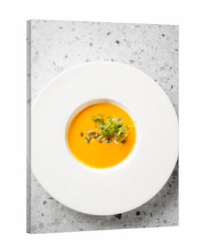 A close-up shot of vibrant orange pumpkin soup, elegantly garnished with green sprouts and pumpkin seeds, served in a white bowl. Appetizing and healthy dish, top view