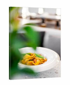 A close-up, selective focus shot of a gourmet pappardelle pasta dish in a white bowl, garnished with fresh basil leaves and served in a modern restaurant.