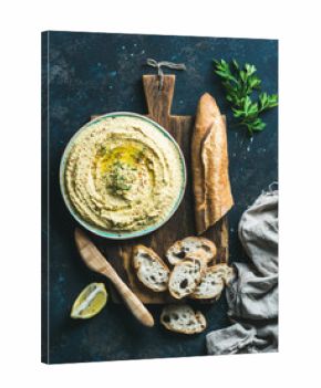 Homemade hummus in blue bowl with lemon, herbs and fresh baguette on rustic wooden serving board over dark blue plywood background, top view, Vegetarian food concept