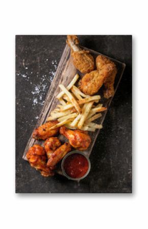 Fast food fried crispy and spicy chicken legs, wings and french fries potatoes with salt and ketchup sauce served on wooden serving board over dark texture background. Top view, space for text