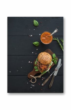Fresh homemade burger on dark serving board with spicy tomato sauce, sea salt and herbs over black wooden background. Top view