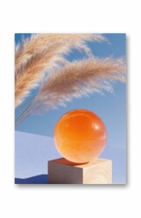 A vibrant orange sphere sits atop a light wood cube,  against a pale blue backdrop, with light tan pampas grass