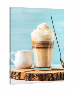 Latte macchiato with whipped cream, serving silver spoon and white pitcher on wooden round board over blue painted wall background