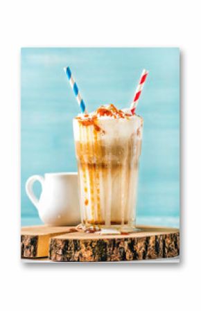 Latte macchiato with whipped cream and caramel sauce in tall glass with straws served with pitcher on wooden board over blue painted wall background, selective focus, vertical composition