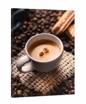       Cup of coffee in the middle of coffee beans with biscuits and tablecloth. Grained product. Hot drink. Close up. Harvesting. Natural background. Energy. Fresh espresso with beans on the foam.