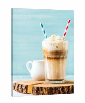 Latte macchiato with whipped cream in tall glass with two straws and pitcher on wooden board over blue painted wall background, selective focus, copy space, vertical composition